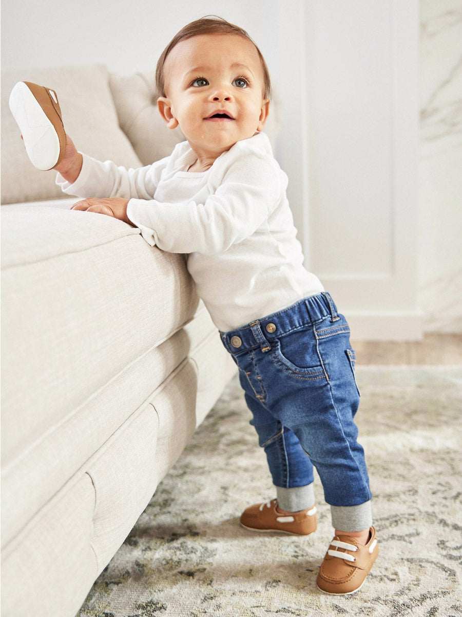 Baby standing on a rug next to a white couch wearing a white sweater, blue jeans, and brown shoes.