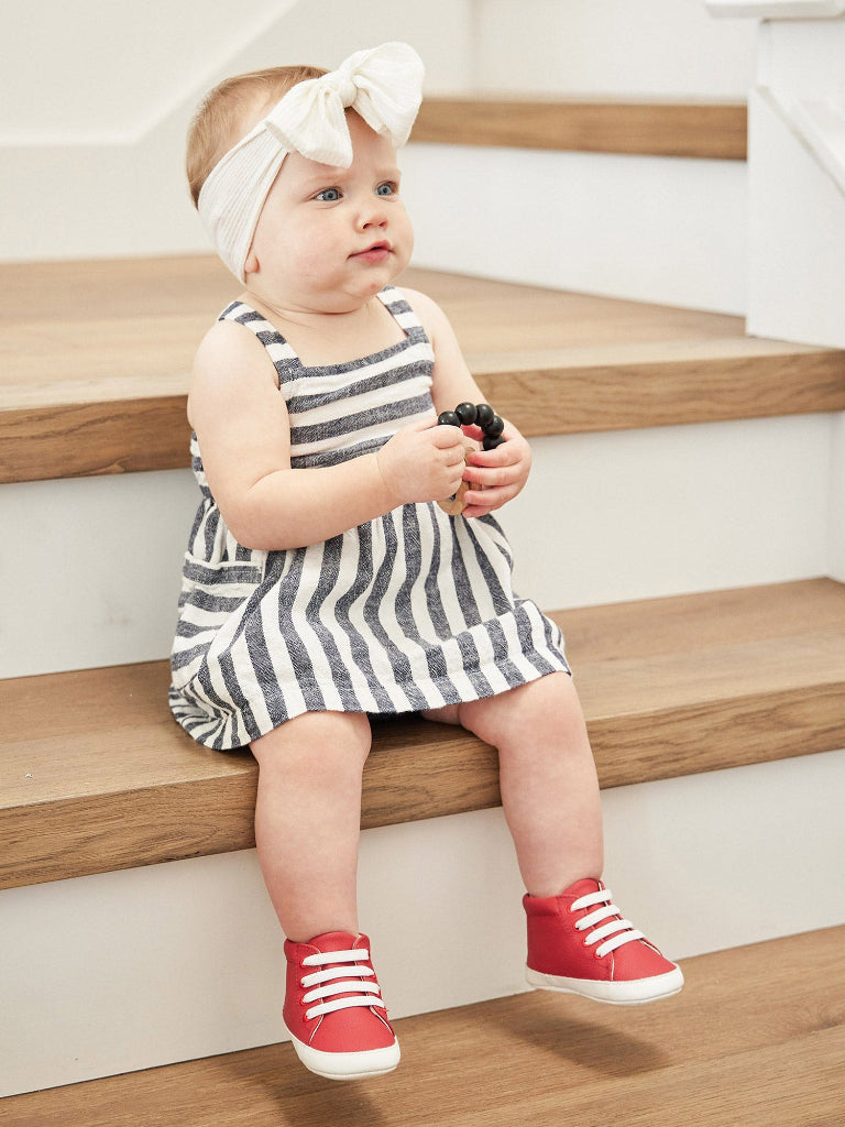 Child wearing a striped dress and red shoes sitting on wooden stairs.