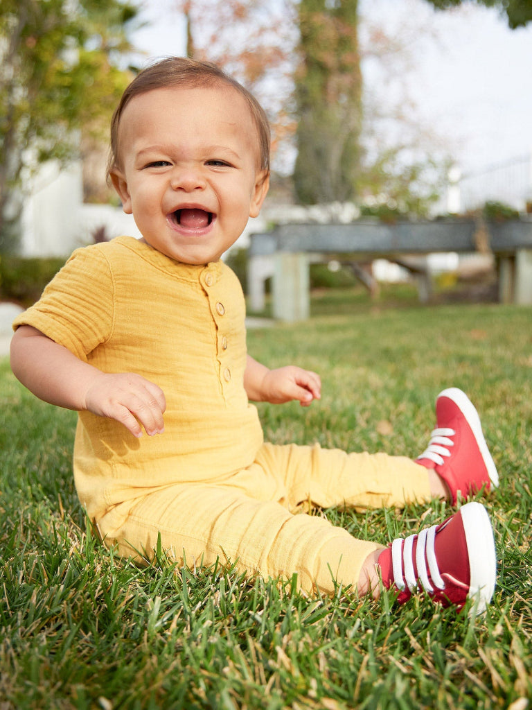 Baby in a yellow outfit with red shoes sitting on grass outdoors