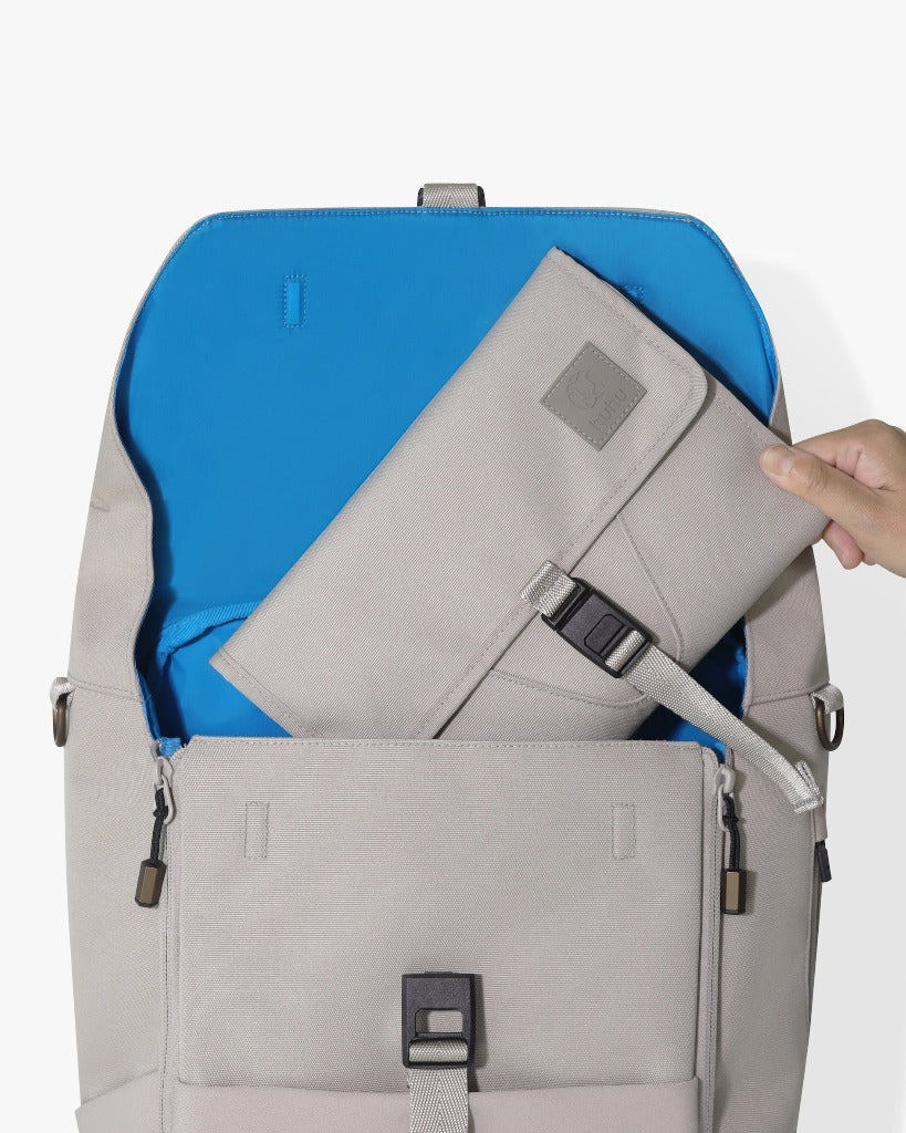 Picture of an open taupe 2.0 backpack with a light-skinned hand placing the taupe changing wallet inside