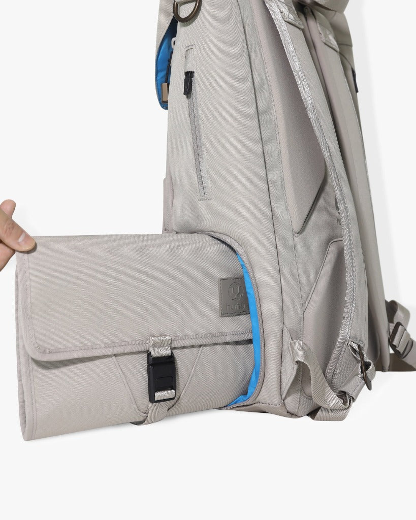 Picture of a side view of the taupe backback against a white background with a light-skinned hand removing the side locker pocket from the bag