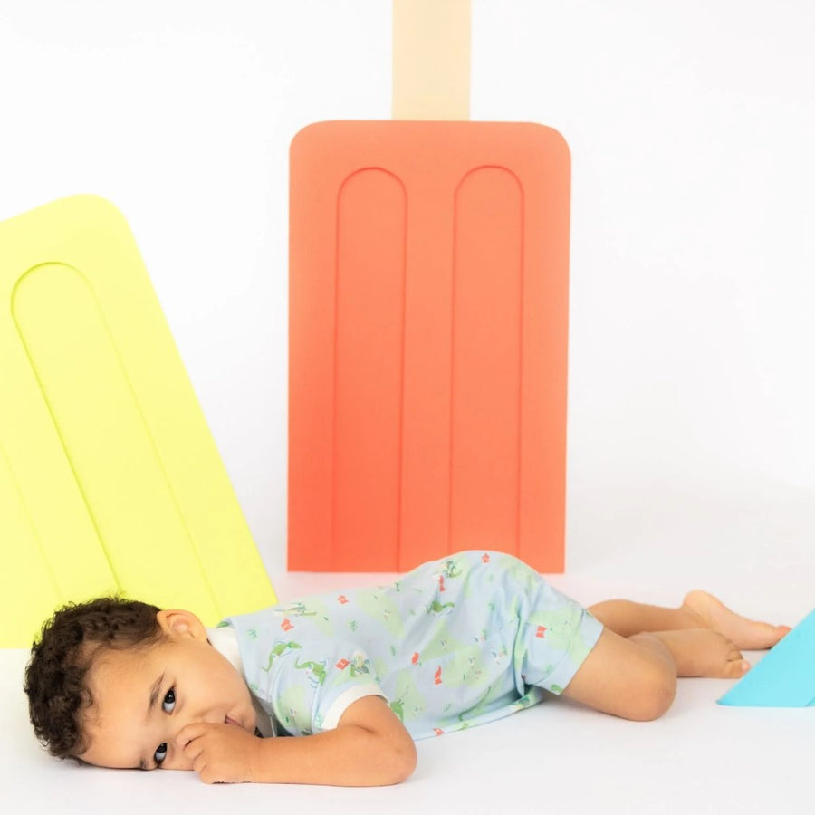 Picture of a baby with a medium skin tone and short dark curly hair, lying on their stomach on a white floor with their thumb in their mouth and looking at the camera. They are wearing the A Putt Above romper. In the background are a coral and a chartreuse colored oversized popsicle decoration