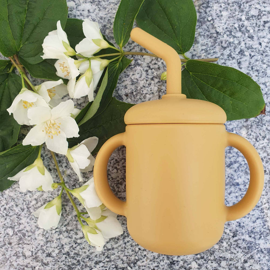 Yellow sippy cup with a lid on a gray surface with green leaves and white flowers.
