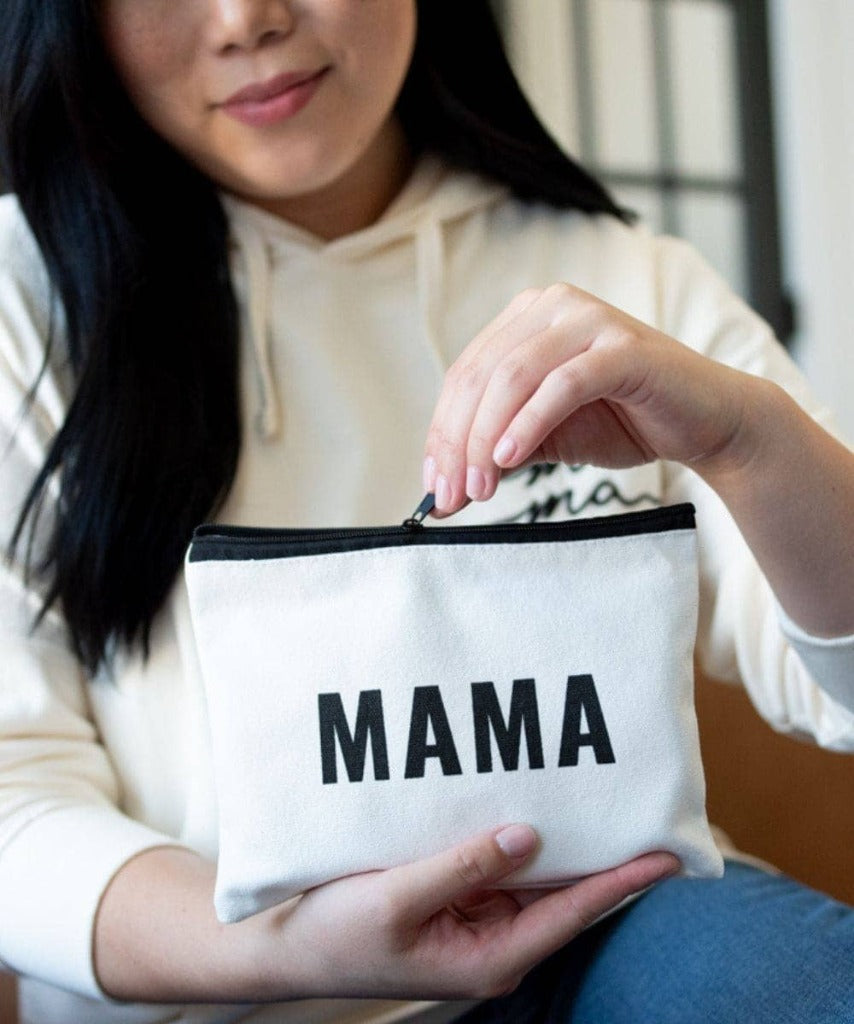 Image shows a person with a medium light skin tone and long dark hair, wearing a cream colored sweatshirt and blue jeans, holding the MAMA pouch and unzipping it