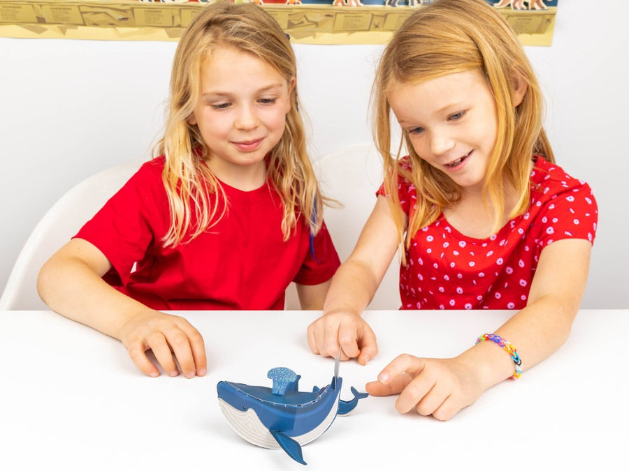 Image shows two children with long dark blonde hair and pale skin tones both wearing red and sitting at a white table and looking at the assembled blue whale and calf which are resting on the table
