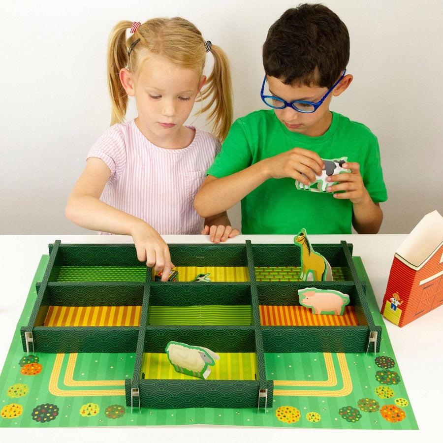 Image of two young children playing with the farmset on a white table. On the left is a child with pale skin, long blonde hair in pigtails, and a pink and white striped shirt, and on the left is a child with light skin, short dark hair, blue glasses, and a bright green t-shirt.
