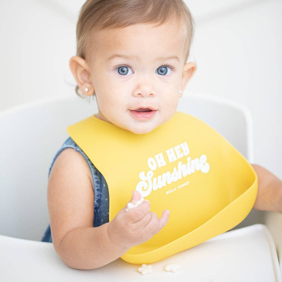 Baby wearing a yellow bib with text, sitting in a high chair.