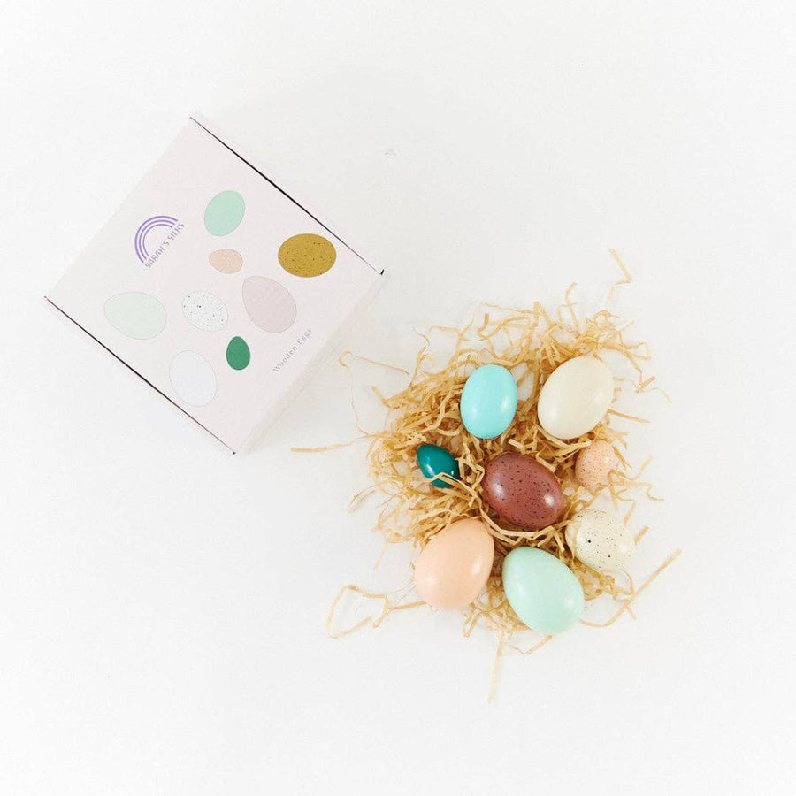 set of wooden painted eggs of various sizes and colors in a nest of paper straw next to their packaging box