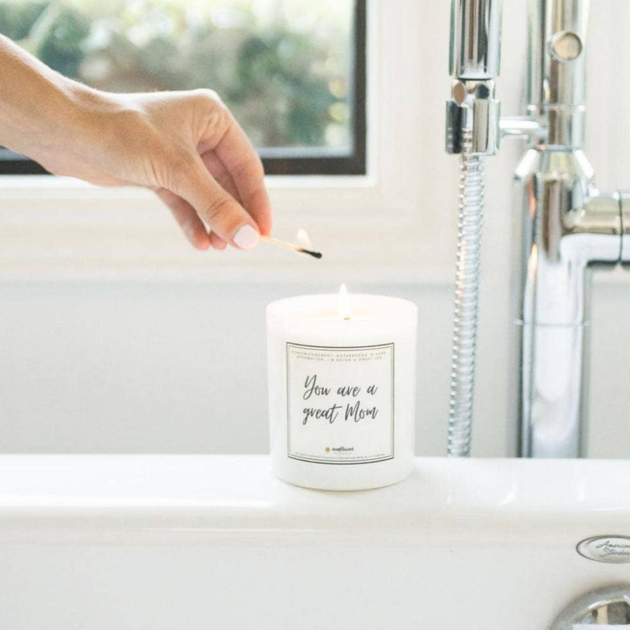 Image shows the Great Mom candle on the edge of a white bathtub, with a person's hand with a light skin tone and white painted nails lighting it with a wooden match
