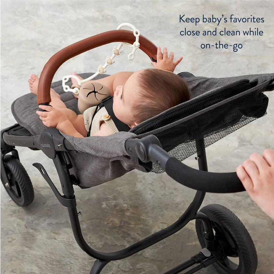 Baby in a stroller with a toy hanging from the handle, on a neutral background.
