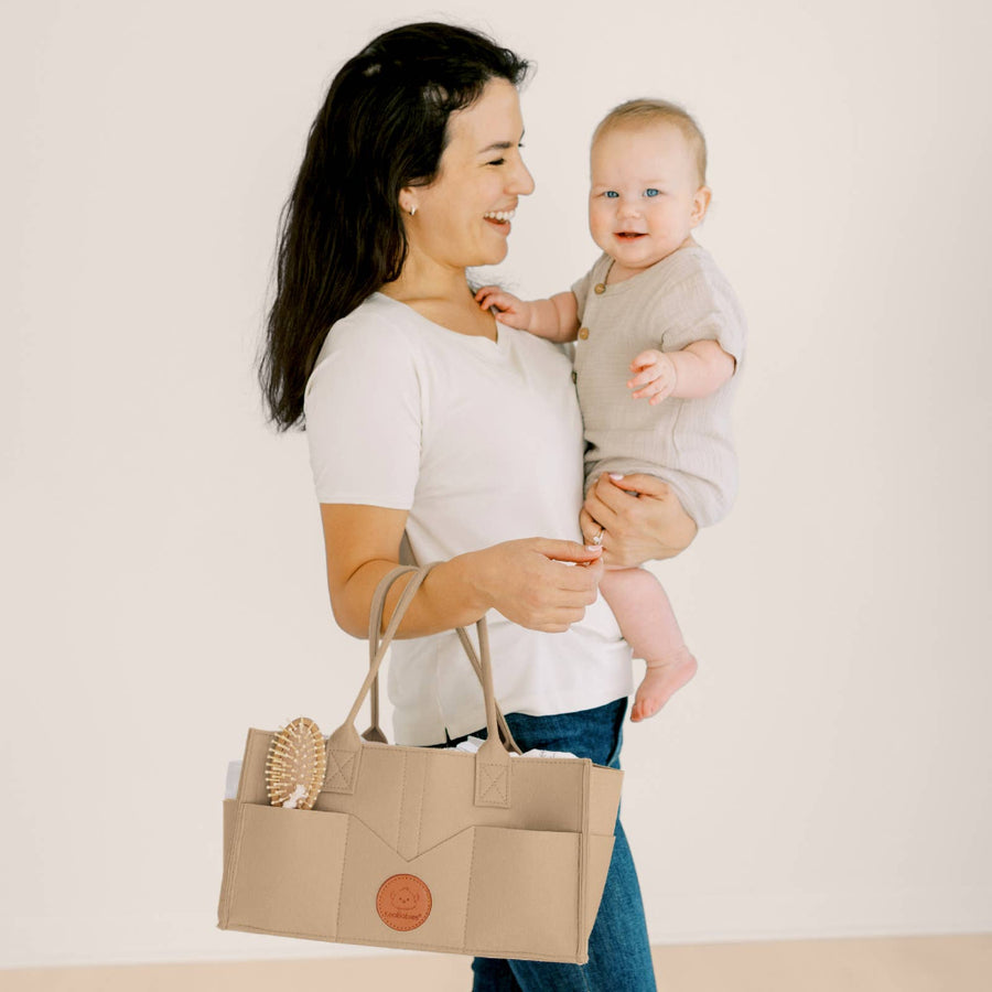 Woman holding a baby and a beige caddy bag with a brown logo on a light background
