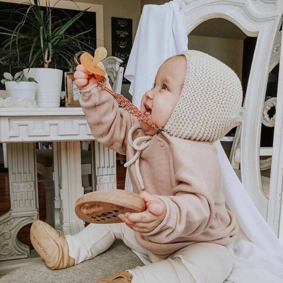 Image of a seated baby with a pale skin tone holding up an ecopacifier. They are wearing tan leather booties, white leggings, a light pink long sleeved top, and an off-white crocheted bonnet. In the background is a white table with potted plants and framed pictures and a large white mirror.