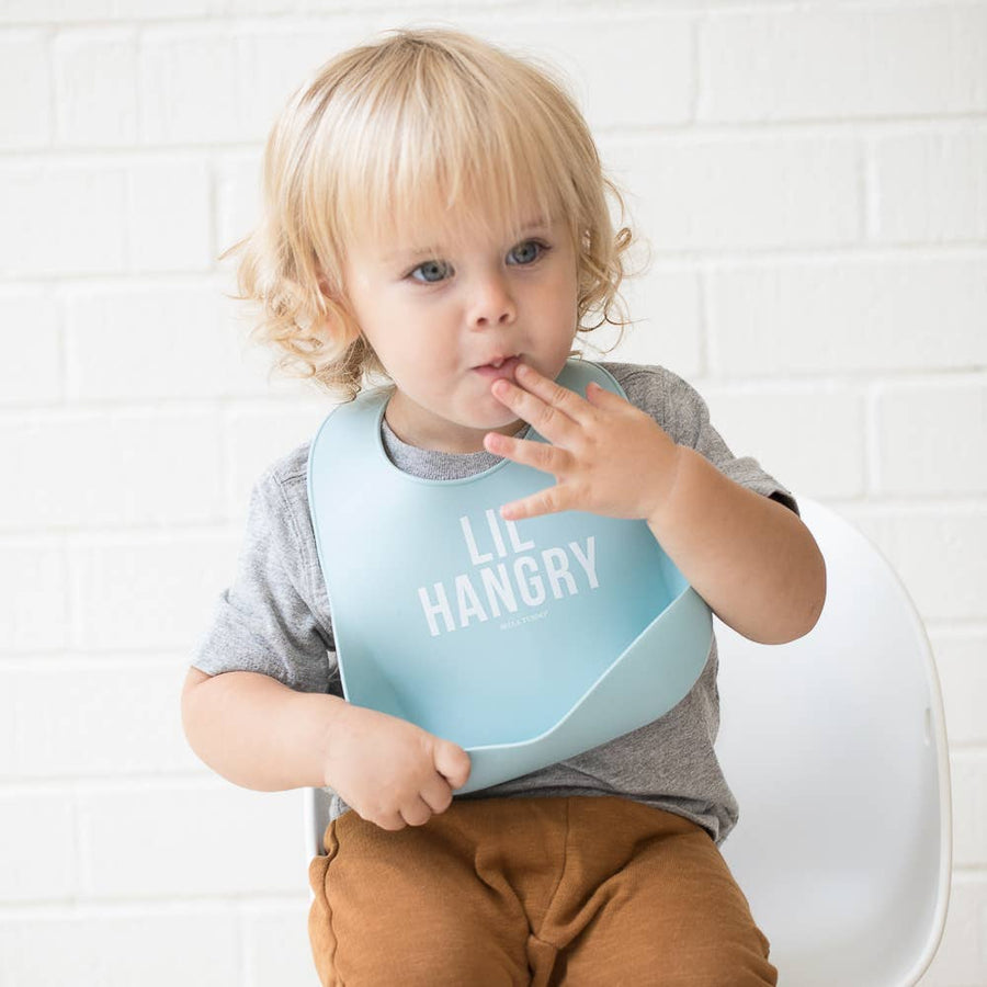 Child wearing a bib with 'LIL HANGRY' text, sitting on a white chair against a white tiled wall.