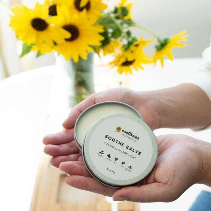 Image shows the salve tin with the lid ajar, being held in a person's upturned hands with a light skin tone, with a white table and a bouquet of sunflowers in the background