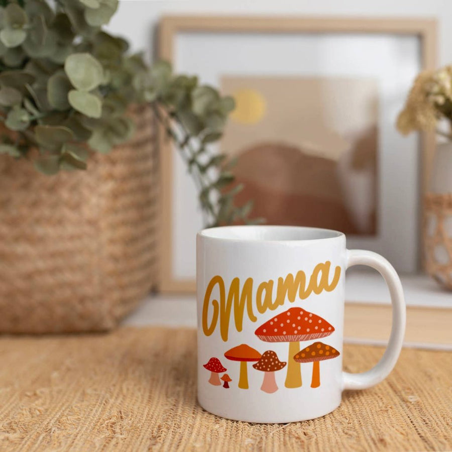 Picture of the mug sitting on a light brown surface that may be a bamboo mat. Out of focus in the background is a plant in a square basket and a framed picture.