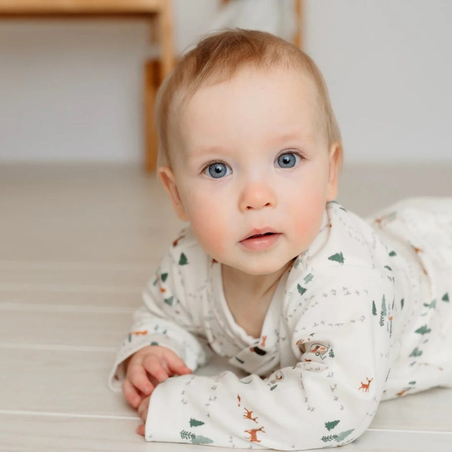Baby wearing a white onesie with animal patterns on a light wooden floor.