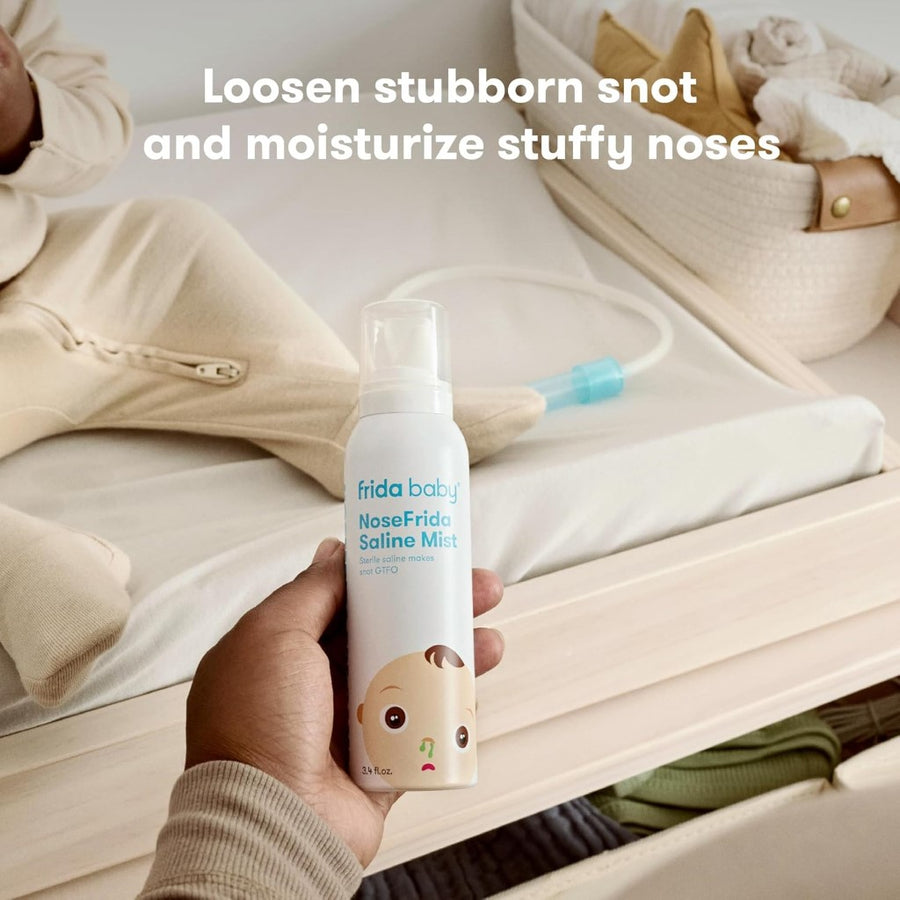 Image of a hand with a medium skin tone holding a can of the NoseFrida Saline Mist in front of a neutral colored changing table, with a seated baby in a beige footie mostly out of frame, a NoseFrida on the changing pad, and a cream basket. Superimposed text reads, 