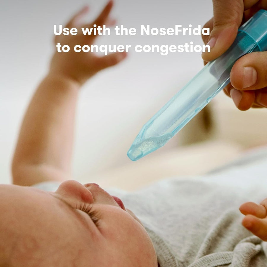 Image of a light skinned baby in a gray onesie, lying on their back with head and shoulders in frame, with a hand with a medium skin tone holding a NoseFrida nasal aspirator above them ready to use. Superimposed text reads, 