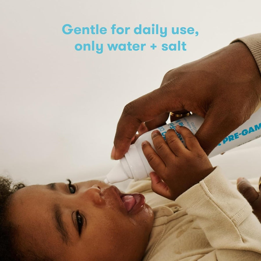 Image of a baby with medium-dark skin and curly dark hair, from the waist up, wearing a cream colored long sleeved outfit and lying on their back, with a hand with a medium-dark skin tone holding the NoseFrida Saline Mist can above them ready to use, against a cream background. Superimposed text reads, 