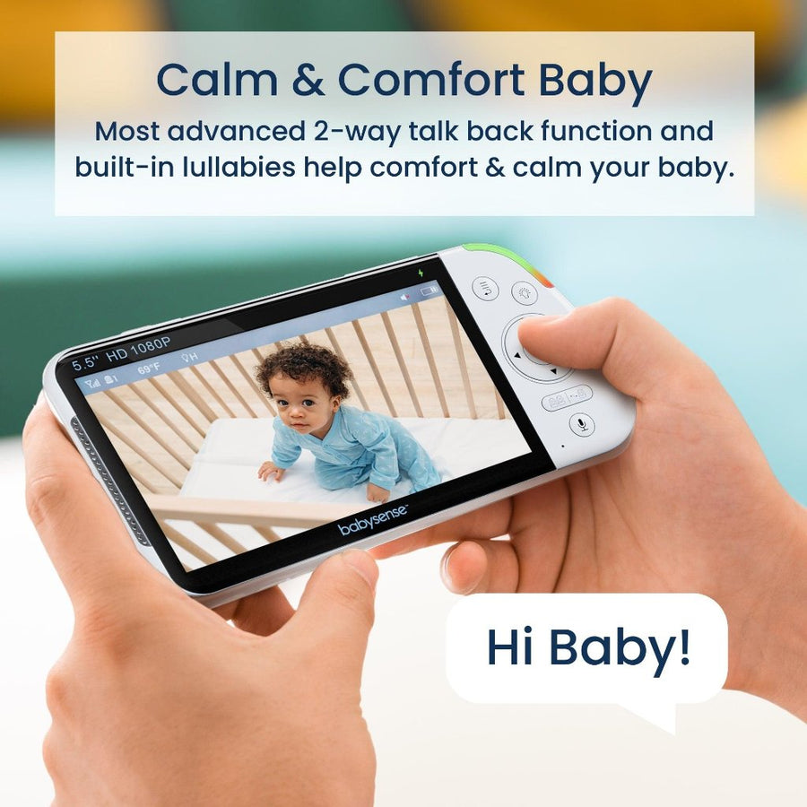 Image shows a pair of hands with a pale skin tone holding the monitor display. On the display screen is a baby with dark curly hair and a medium skin tone wearing a light blue footie, sitting in a crib and looking up at the camera. Text at the top of the image reads, 