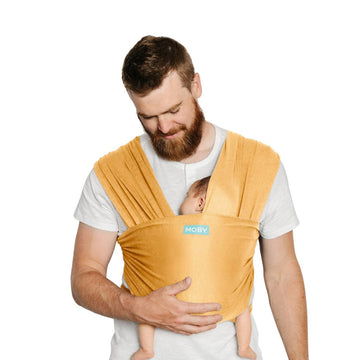 Picture of a light-skinned man with short reddish brown hair, moustache, and beard, wearing a Marigold Moby Classic Wrap carrier over a white t-shirt. He is looking down at the sleeping baby in the carrier with one hand on baby's bottom