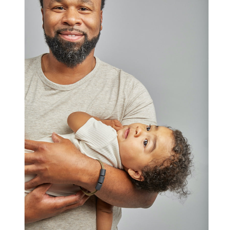 A smiling man is wearing the Saddle Adventure Teething bracelet and holding a child in his arms. He has short hair, a medium-dark skin tone, and a beard and is wearing a plain t-shirt. The child has a medium skin tone and curly hair and is laying back across his arms while wearing a light-colored ribbed top.