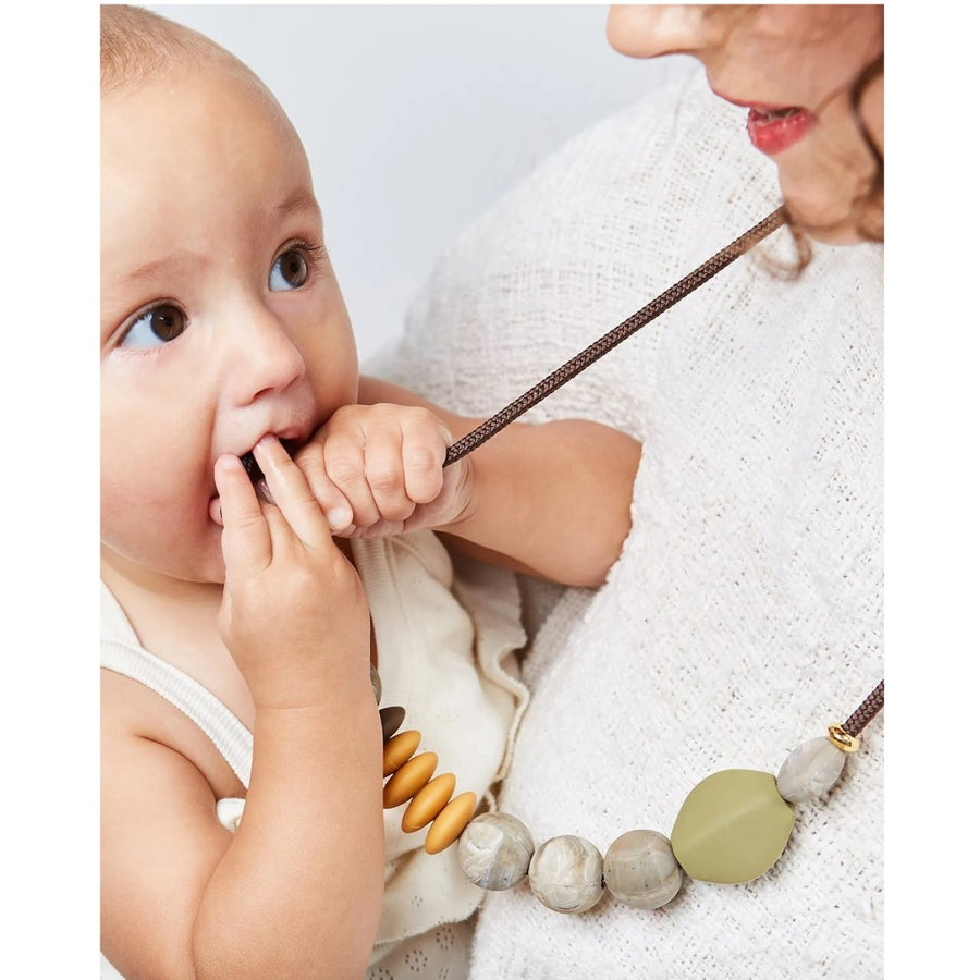 The image shows a baby in a person's arms, chewing on the Saddle Signature Teething Necklace which is being worn by the adult. They are both wearing off white clothing. The baby has brown eyes and a light skin tone and the adult has brown curly hair and dark pink lipstick