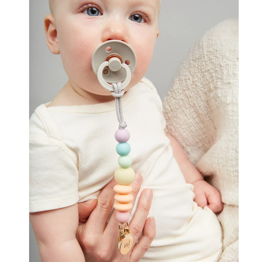 A baby is in a person's arms and is using a pacifier that is attached to the Rainbow Sherbet pacifier clip
