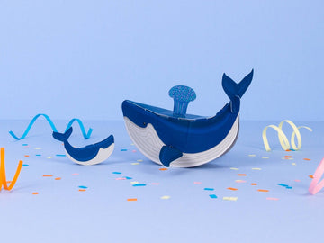 Image shows the assembled whale and calf sitting on a blue surface with multi colored confetti and paper spirals, against a blue background