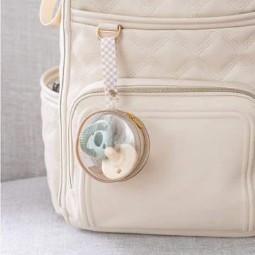 A close-up picture of a pale pink diaper bag sitting on a gray upholstered surface with the Itzy clear paci case attached to a bag loop with two pacifiers inside