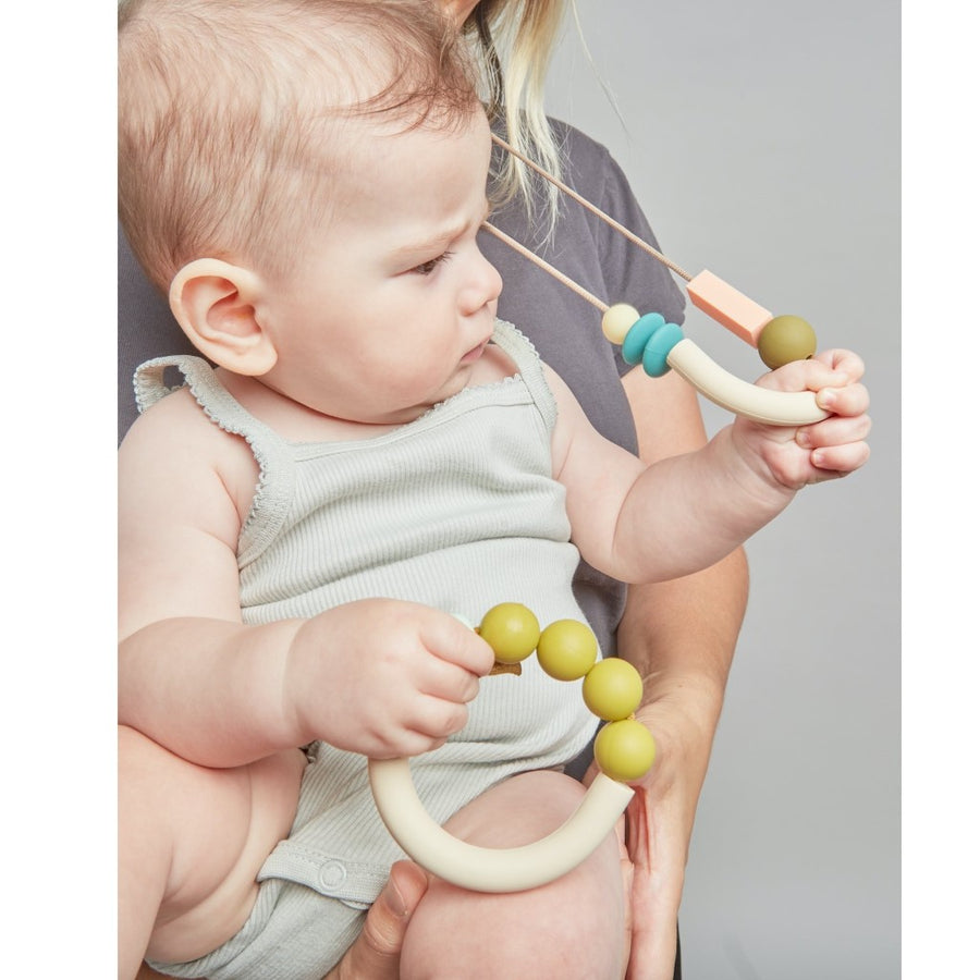A baby in a person's arms is holding the Dewdrop Arch teething bracelet and playing with the Dewdrop Balance necklace that the person is wearing