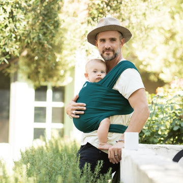 Picture of a man wearing a dark teal colored Moby Classic baby wrap with baby, standing outside near plants and a wall