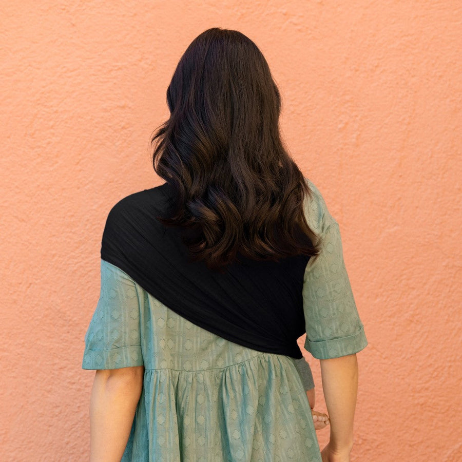 Picture of a woman wearing a Moby Wrap Ring Sling in Onyx, from the back. It shows the fabric spread over her left shoulder and across her back. She has long dark hair and is wearing a blue dress