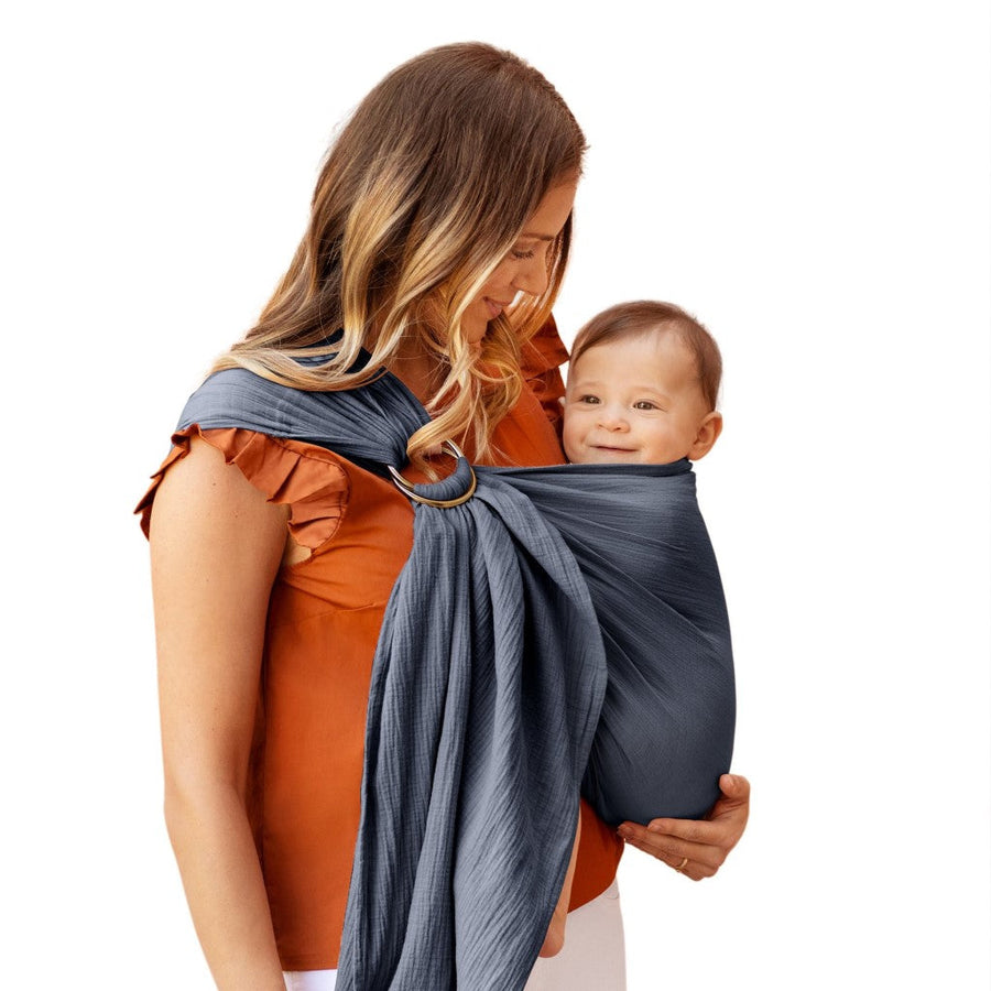 Picture of a woman holding a baby in the Moby Wrap Ring Sling in Flint. She has long dirty blond hair and a light skin tone and is wearing a short sleeved orange top with the gray sling over her right shoulder and looking down at a light skinned baby with short brown hair sitting upright and smiling