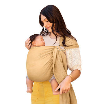 Picture of a woman holding a baby in a Moby Wrap Ring Sling in Saffron. She has long dark hair and a medium skin tone and is wearing a cream colored top with lace half sleeves and yellow pants, and looking down at a sleeping baby with short dark hair and a medium skin tone in a legs-out position in a pale yellow ring sling carrier