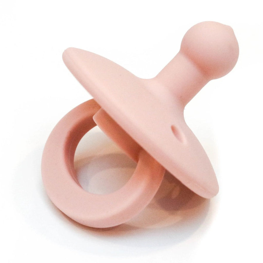 Picture of an OLI2 pacifier in a peachy pink color. It has a loop handle, a round flange with several holes, and a bulb-shaped nipple